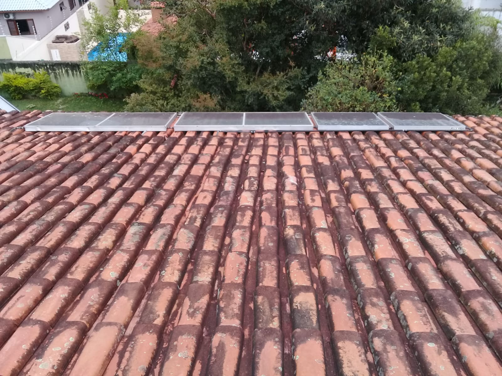 Telhado residencial com contraste visível entre telhas já lavadas (cor laranja viva) e áreas ainda sujas de musgo/fuligem, com painéis solares ao fundo